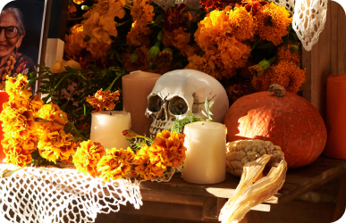 An ofrenda with marigolds, lit candles, a decorative skull and a pumpkin, a framed photograph of an older woman resting at the left.