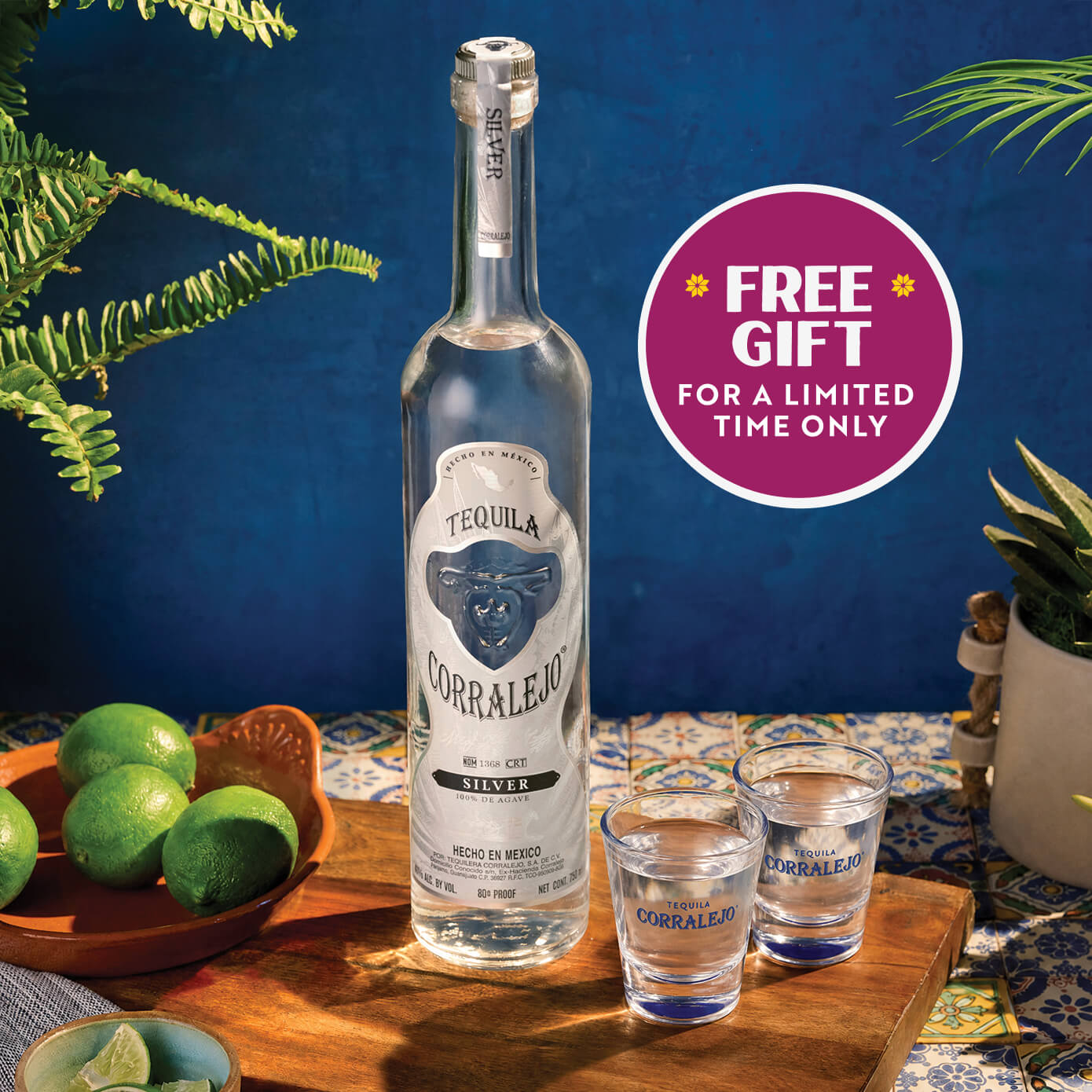 A bottle of Corralejo Silver tequila stands on a wooden tray beside two matching Corralejo shot glasses, with limes, plants, and a blue patterned backdrop. A magenta circular badge reads 'FREE GIFT FOR A LIMITED TIME ONLY.
