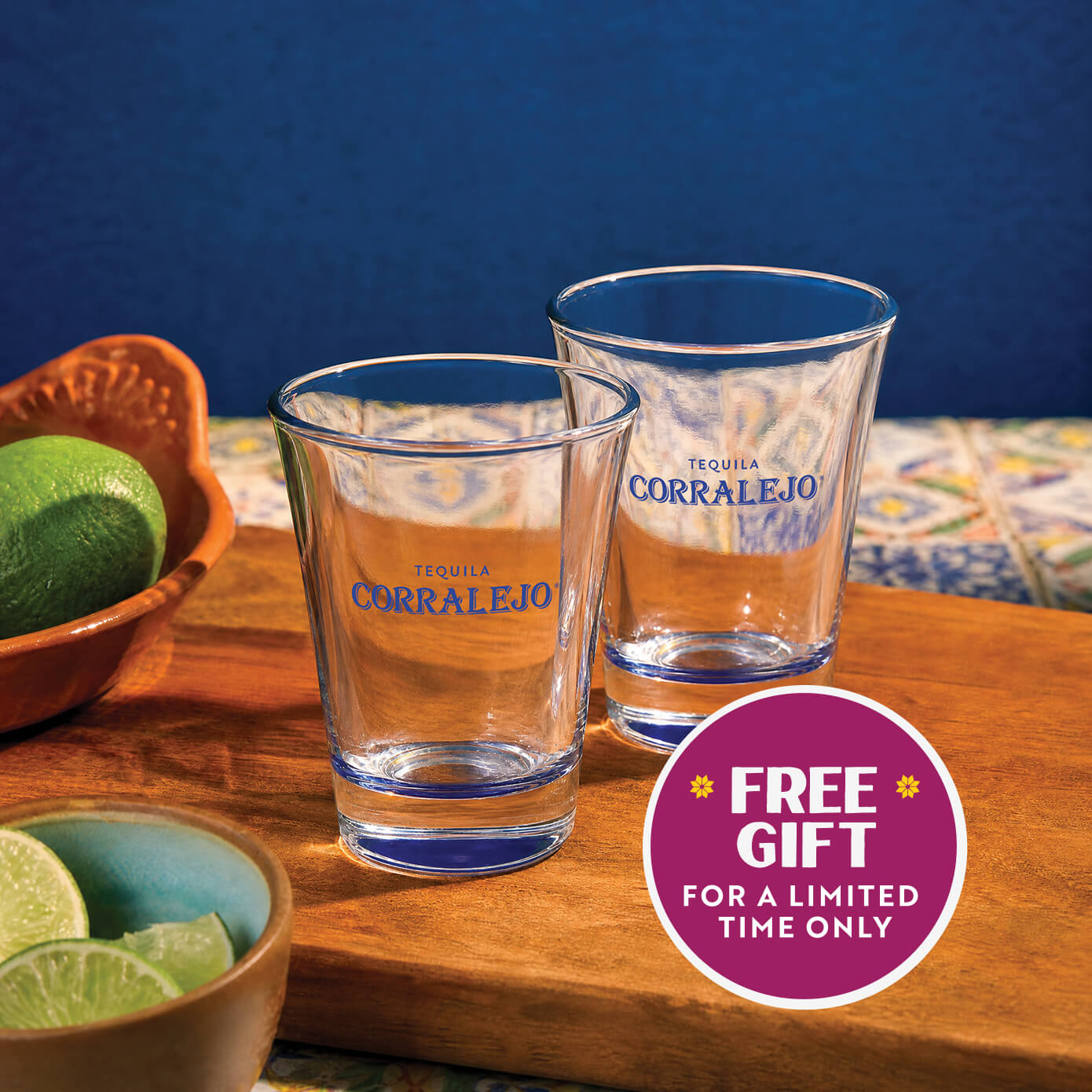 Two clear Corralejo Tequila shot glasses with blue lettering and blue rim accents, displayed on a wooden board with limes and bowls against a blue background. A magenta promotional badge reads 'FREE GIFT FOR A LIMITED TIME ONLY'.