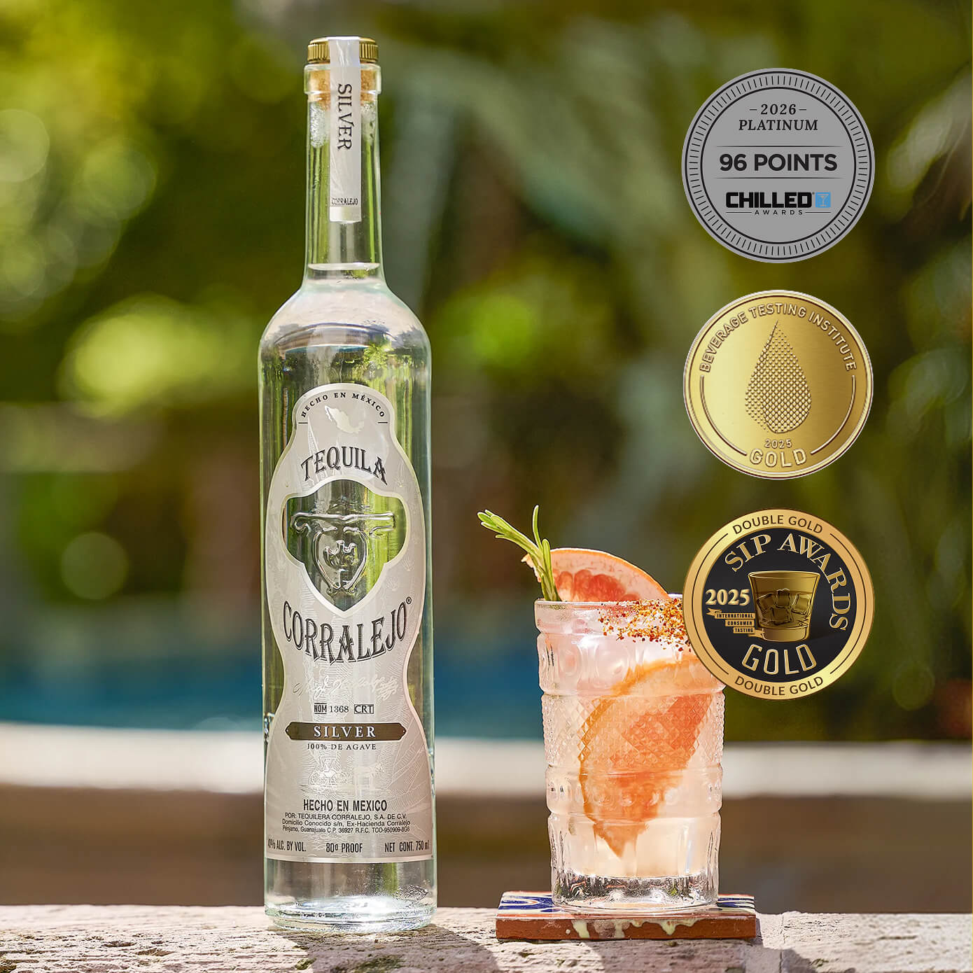 A bottle of Corralejo Silver Tequila stands next to a glass of mixed drink garnished with grapefruit, with three award seals on the right against a blurred outdoor background.